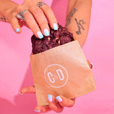Hand holding a Red Velvet New York Cookie with white chocolate chips partially in a Crumbs & Doilies bag against pink background